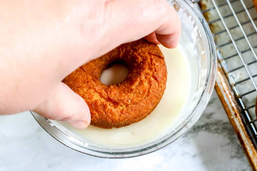 a hand dipping a donut into a bowl of glaze