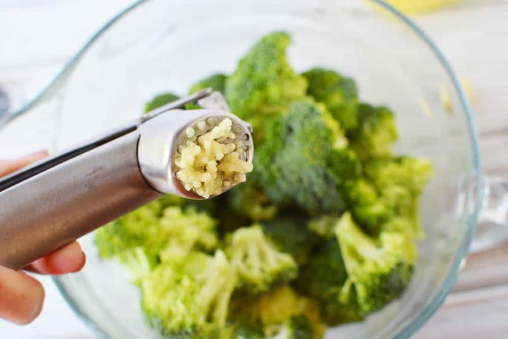 garlic in a garlic press over a bowl of broccoli 